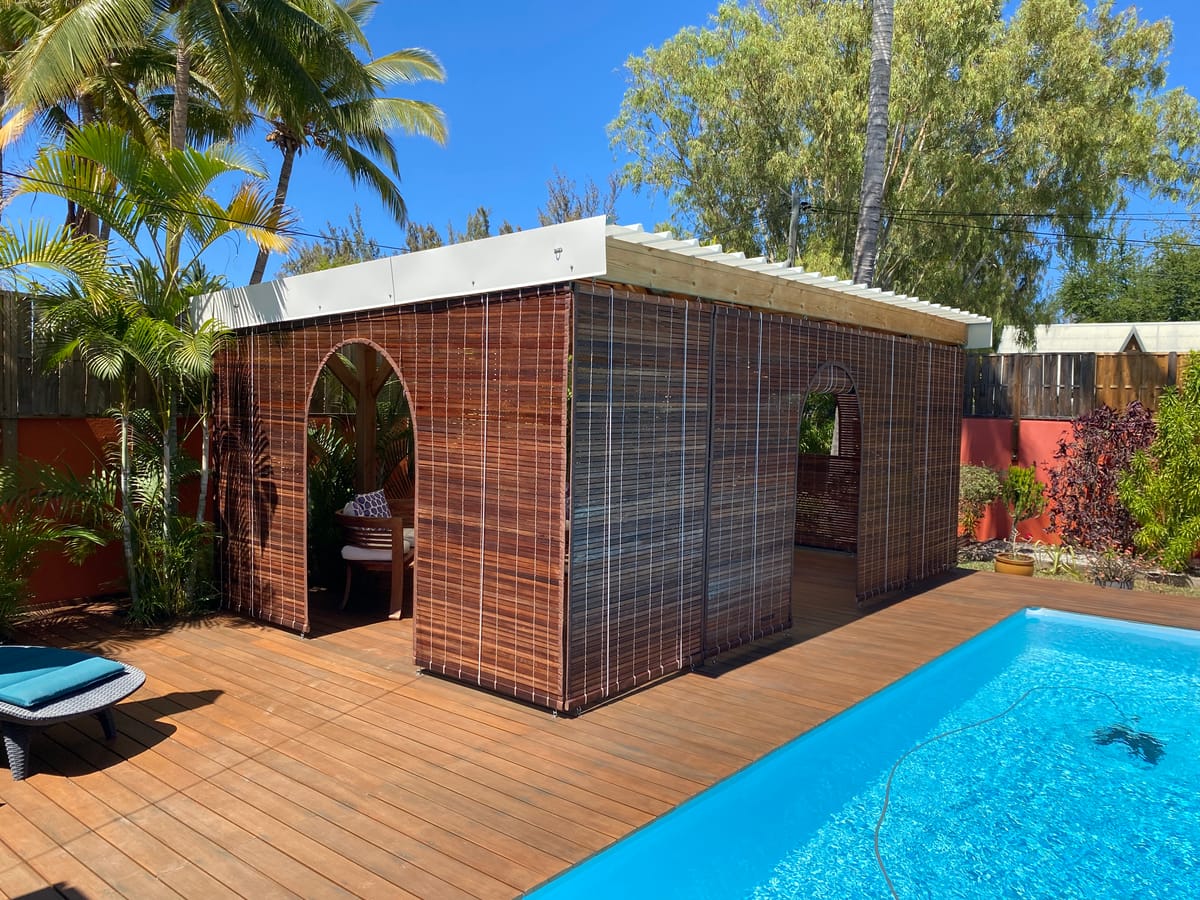 Kiosque de jardin habillé de stores mauriciens en bois avec ouvertures en arche, au bord d’une piscine