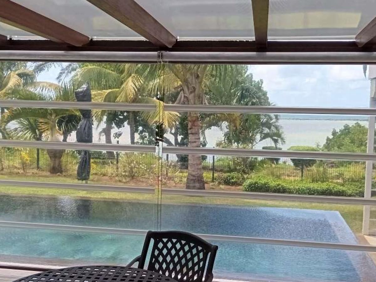 Store vertical guidé transparent baissé sous une pergola, vue dégagée sur la piscine et le jardin